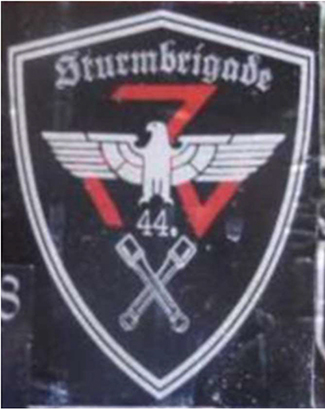Wappenschild mit weißem Schriftzug in gebrochener Schrift „Sturmbrigade“ mit Reichsadler, der Zahl „44.“, gekreuzten Stielhandgranaten sowie einer roten Triskele im Hintergrund