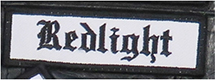 Dieses weiße Kennzeichen mit schwarzer Umrandung in Form eines Rechtecks enthält die Aufschrift „Redlight“, übersetzt Rotlicht, in schwarzen Buchstaben. Das Kennzeichen unterstreicht die Nähe des Trägers zum Rotlichtmilieu.