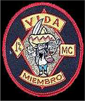 Dieses schwarze Kennzeichen mit roter Umrandung in Form eines Kreises, enthält einen nach vorne blickenden dickbäuchigen Mann mit Sombrero, in dem sich zwei Einschusslöcher befinden. Pistole und Schwert hält er in den Händen. Ein nach obenhin gerundeter, gelber Bogen mit roter Umrandung enthält das spanische Wort „VIDA“ in roten Großbuchstaben, übersetzt Leben. Ein gelber, nach untenhin gerundeter Bogen enthält das spanische Wort „MIEMBRO“, übersetzt Mitglied. Links des Mannes ist das rotumrandete, gelbe, rautenförmige „1 Prozent Zeichen“ abgebildet. Rechts des Mannes in einem rotumrandeten gelben Viereck, die Abkürzung „MC“.